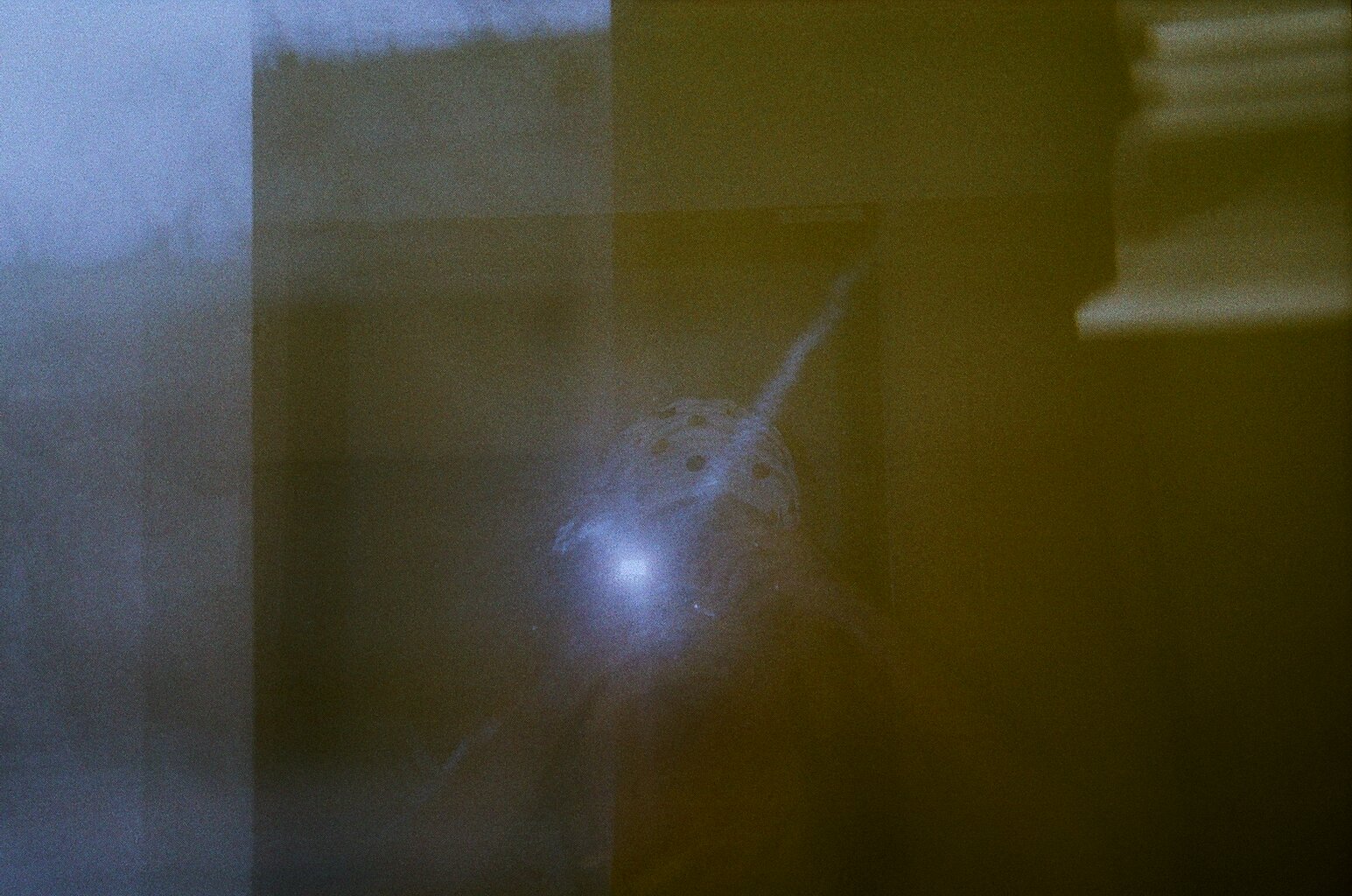 Eine Farbfotografie. Eine Person mit bunter Mütze spiegelt sich undeutlich ine einer Glasscheibe. Die Person fotografiert, das Blitzlicht strahlt hell.