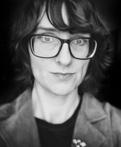 Die Schwarzweißfotografie zeigt Franziska Schiedung. Franziska Schiedung trägt eine Brille. Am Jacket ist ein kleiner Anstecker angebracht.