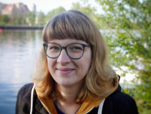 Die Farbfotografie zeigt Stefanie Huhnstock. Sie trägt eine grüne Brille, ein gelbes Hoodie und eine schwarze Jacke. Im Hintergrund ist das Wasser eines Flusses und grüne Blätter eines Baumes.